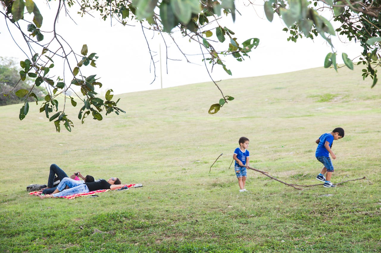 Boys playing with tree twig near unrecognizable lying mother and father on textile in summertime