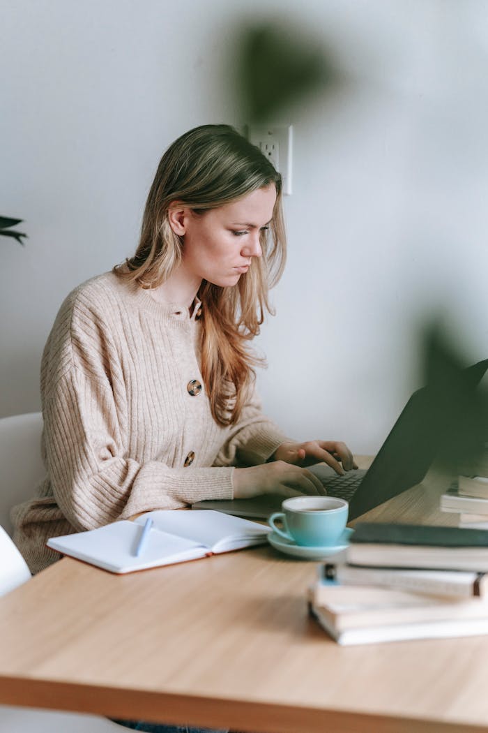 Side view of concentrated female freelancer browsing internet on netbook while sitting at table with notepad and cup of hot drink